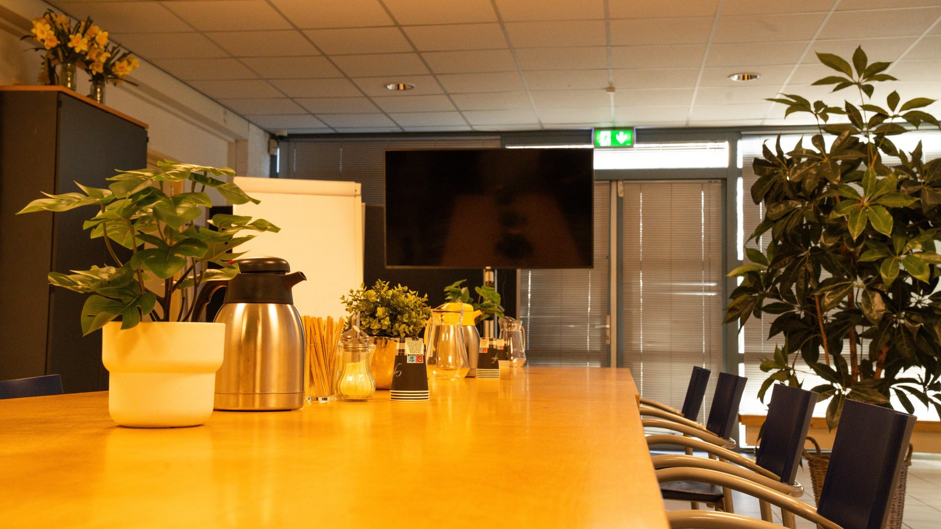 Detail van de vergadertafel: koffiekannen, bekers en planten, met het tv-scherm en de flipover op de achtergrond