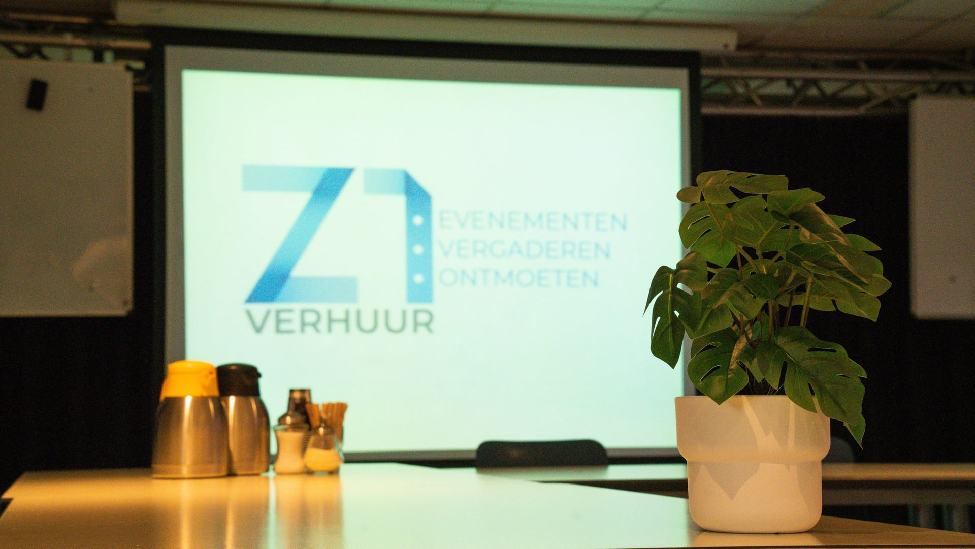 Detail van de tafel in de multifunctionele ruimte: koffiekannen, roerstaafjes en een plant voor het projectiescherm met het logo van Z1 Verhuur