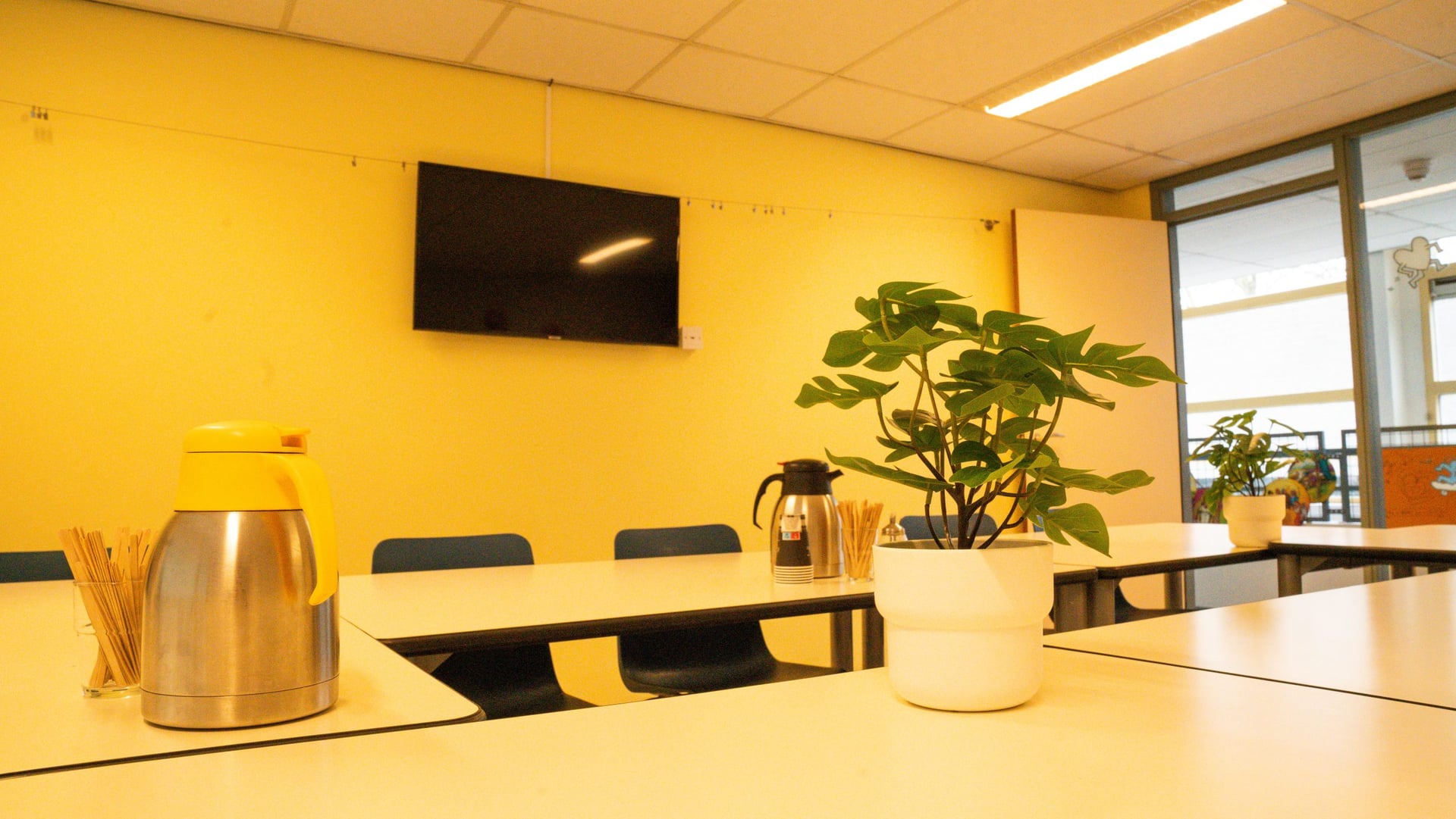 Kleinere vergaderruimte met een lichte wand, tv-scherm aan de muur, koffiekannen en een plant op tafel
