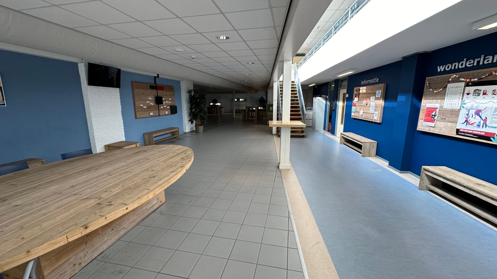 De ontmoetingsruimte met lange houten tafels en banken, uitkijkend op de centrale hal