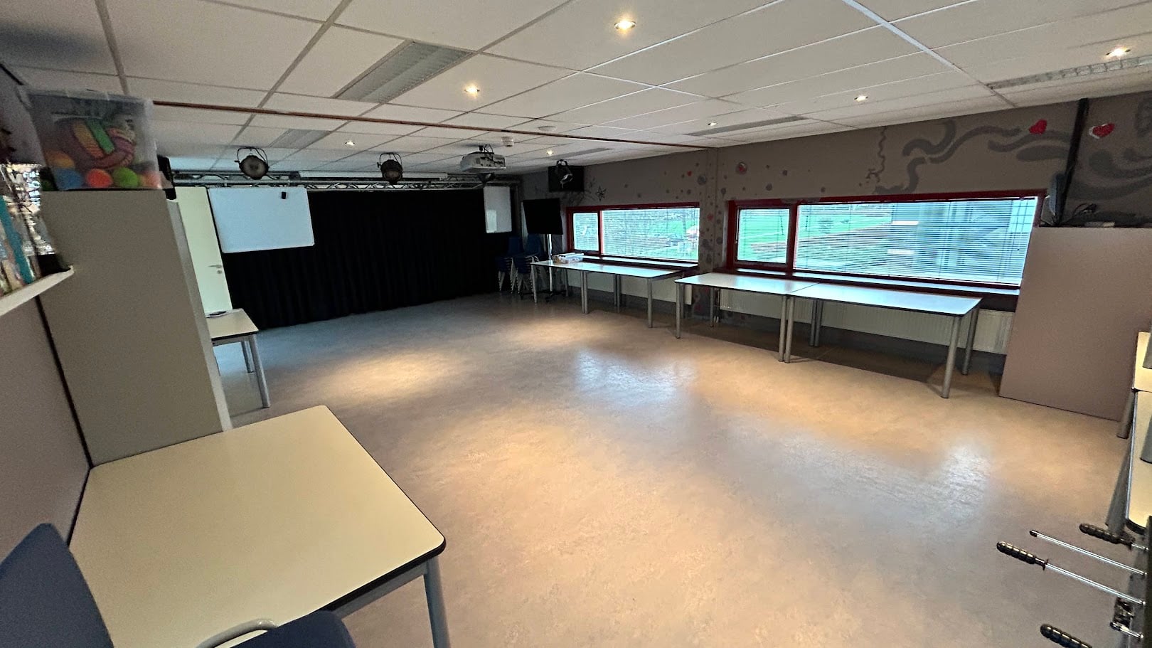 Andere hoek van de multifunctionele ruimte, met projectiescherm, verduisteringsgordijn en daglicht door de ramen